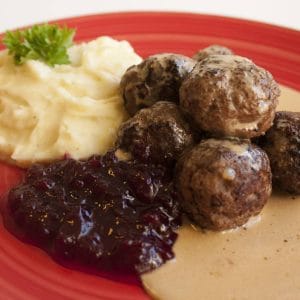En tallrik med klassiska köttbullar, serverade med potatismos och lingonsylt. Köttbullarna är runda och har en gyllene yta, medan potatismoset har en slät konsistens och är garnerat med smör. Lingonsylten är tjock och röd och ligger bredvid köttbullarna på tallriken.